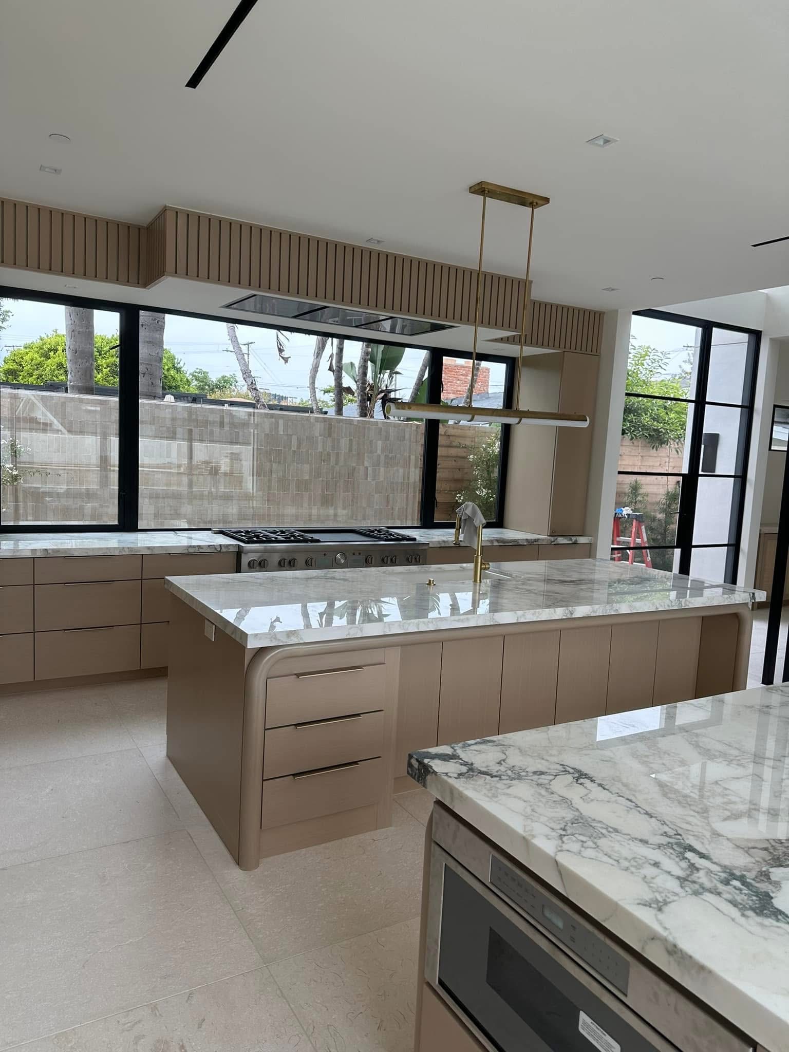 Modern kitchen with marble countertops, tan cabinetry, island with sink, and large windows overlooking outdoor space